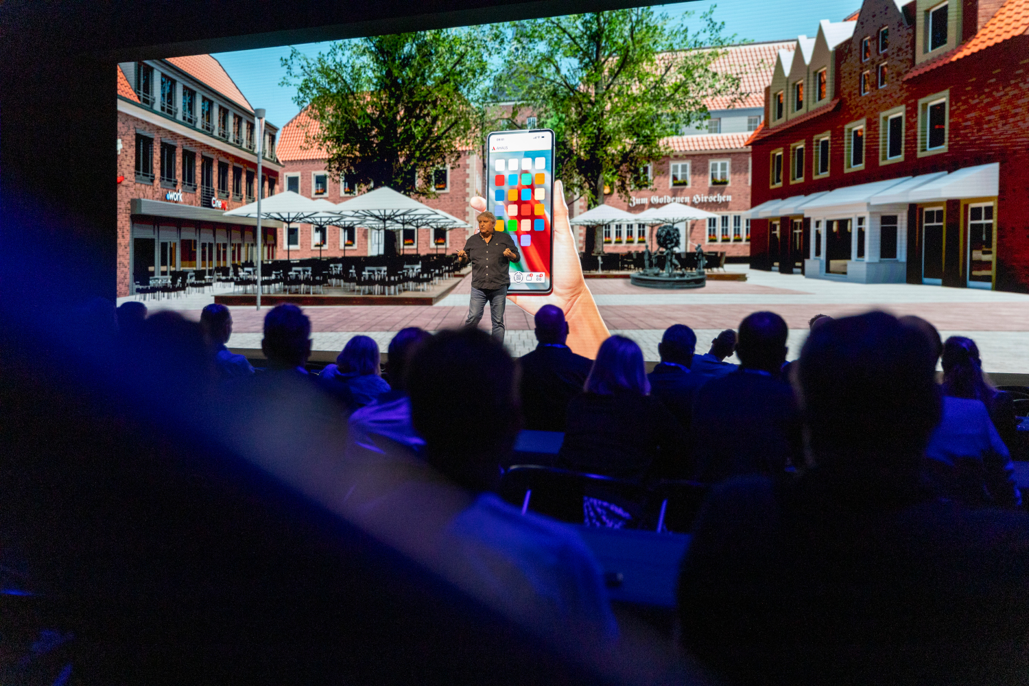 Ein Mann steht auf einer Bühne. Im Hintergrund ist eine Präsentation mit einem Stadtpanorama zu sehen. Man sieht die Zuschauenden von hinten.